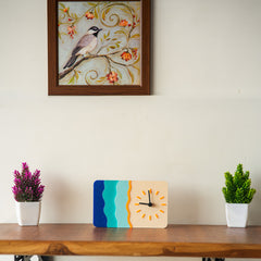 Decorative clock on a wooden shelf with two potted plants and a framed picture of a bird on a branch.