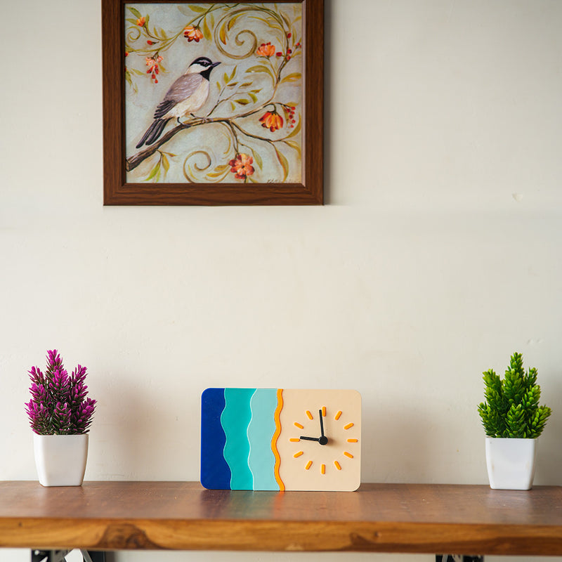 Decorative clock on a wooden shelf with two potted plants and a framed picture of a bird on a branch.