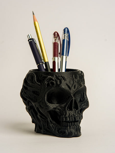 Black skull-shaped pen holder with various pens on a light beige background